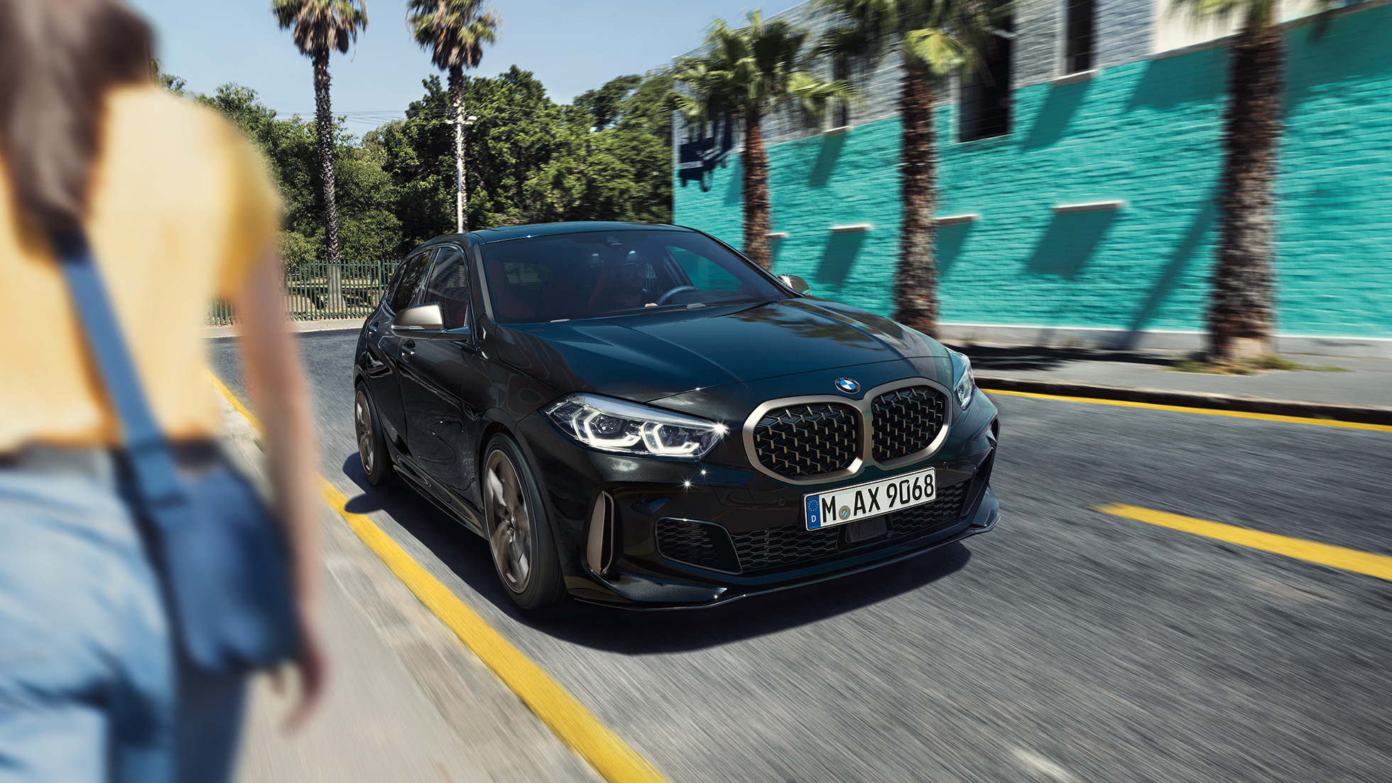 BMW M135i xDrive, vue de trois quarts avant. BMW M135i xDrive en Saphirschwarz metallic, vue de trois quarts avant, avec femme à l’avant-plan.