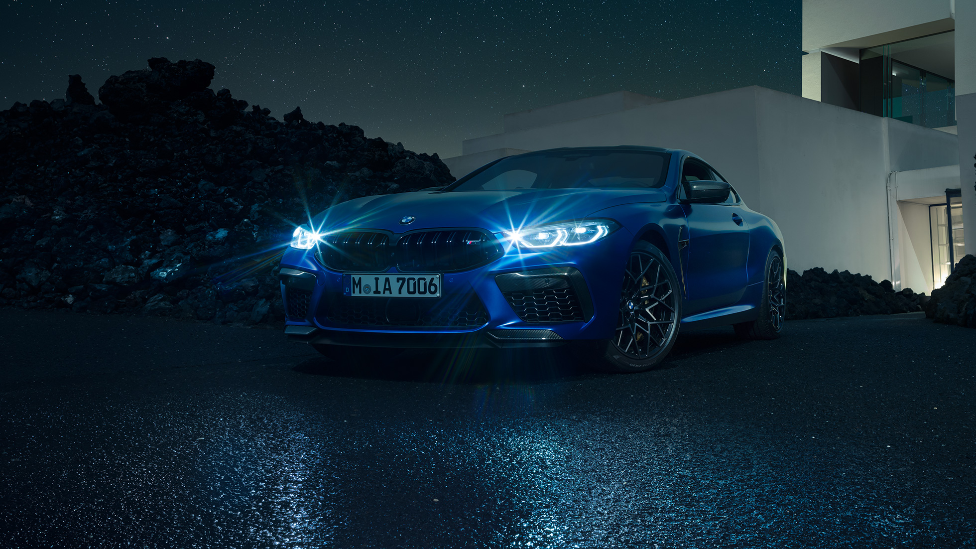 BMW M8 Competition Coupé, vue de trois quarts avant. BMW M8 Competition Coupé en Marina Bay Blau metallic avec phares LED allumés, vue de trois quarts avant à l’arrêt devant une villa, la nuit.