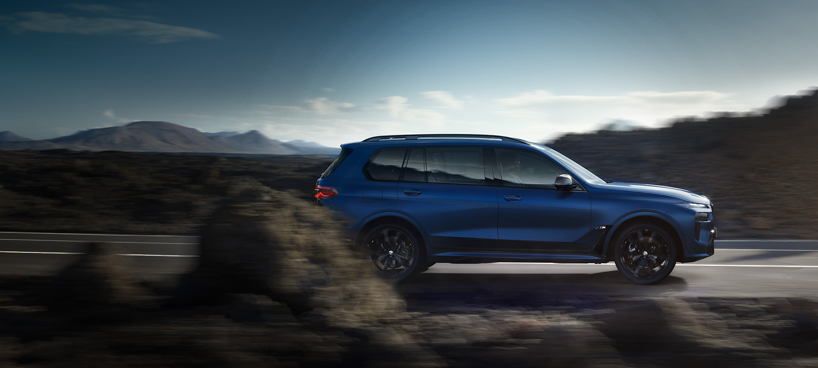 BMW X7 M60i xDrive G07 LCI Frozen Marina Bay Blue vue de profil roulant sur la route BMW X7 M60i xDrive G07 LCI Frozen Marina Bay Blue vue de profil roulant sur la route devant un paysage de montagnes