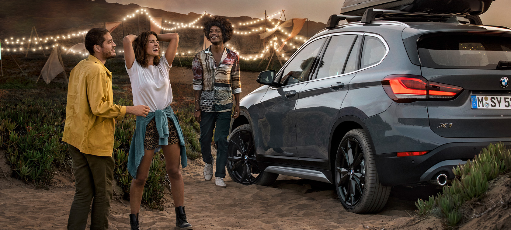 Trio heureux à bord d’une BMW X1 équipée d’accessoires printemps-été BMW Trio heureux à la plage, à bord d’une BMW X1 gris minéral équipée d’accessoires printemps-été BMW, vue latérale de trois quarts