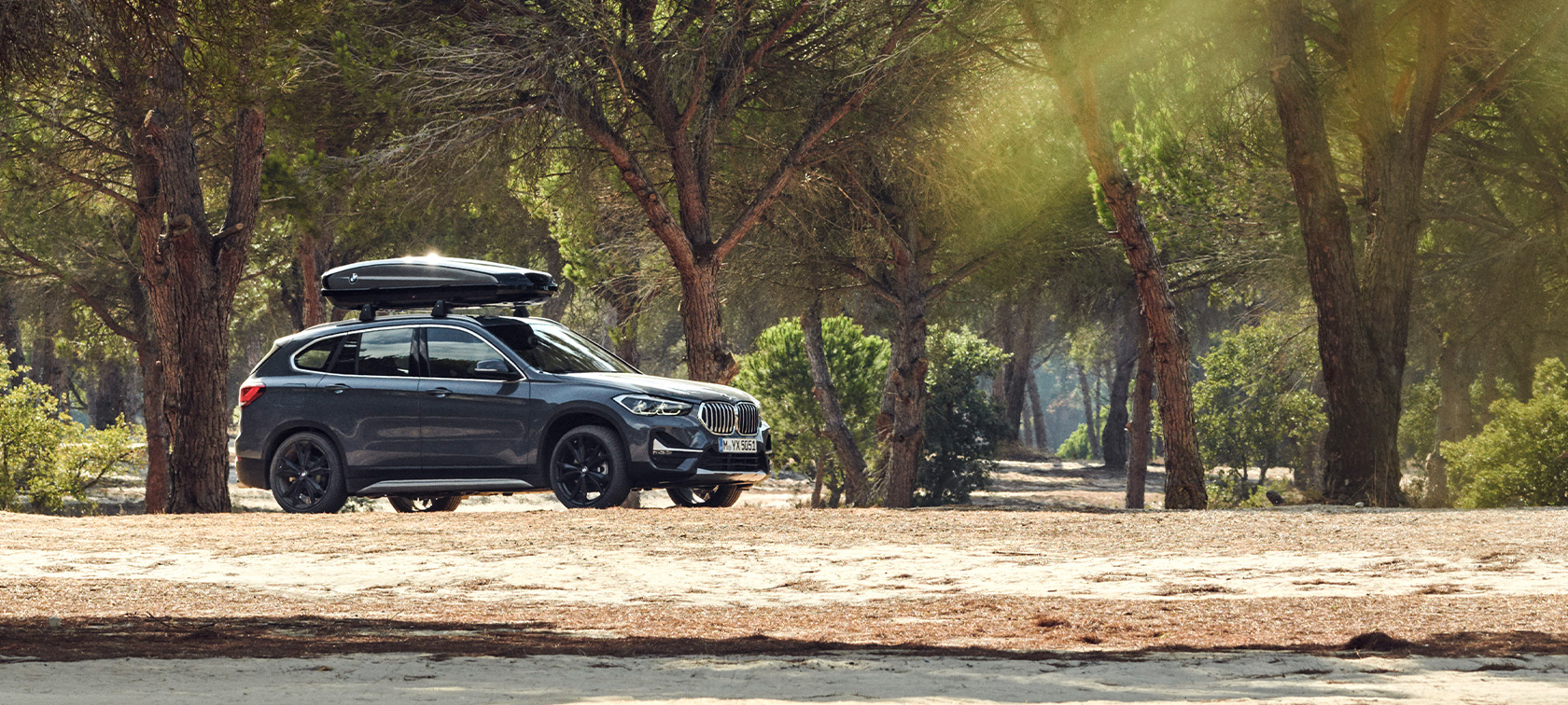 BMW X1 gris minéral équipée du coffre de toit BMW, vue latérale, accessoires printemps-été BMW BMW X1 gris minéral équipée du coffre de toit BMW au milieu de la forêt, vue latérale, accessoires printemps-été BMW