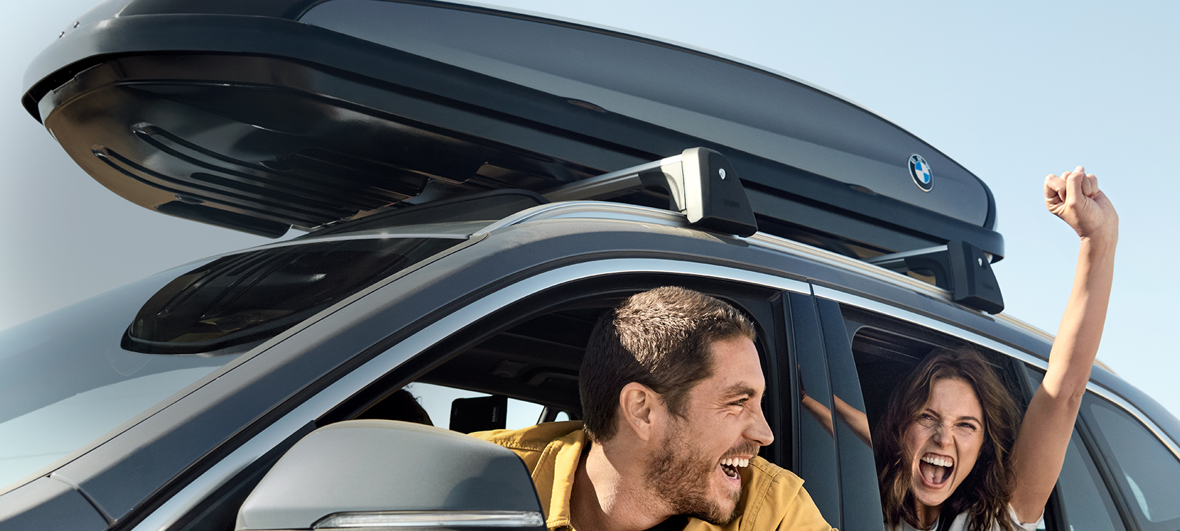 Couple heureux à bord d’une BMW X1 gris minéral équipée d’accessoires printemps-été BMW Couple heureux à bord d’une BMW X1 gris minéral équipée d’accessoires printemps-été BMW sous un ciel d’azur, vue latérale de trois quarts